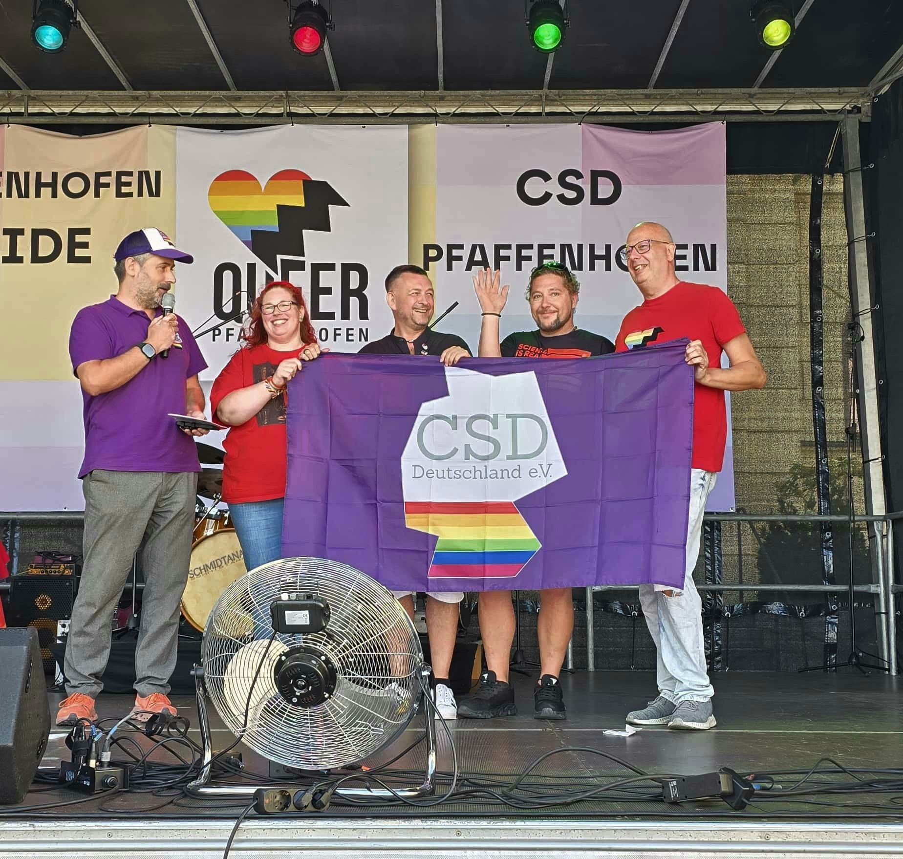 Mehrere Mitwirkende stehen auf der CSD-Bühne und halten gemeinsam ein Banner vor dem Veranstaltungshintergrund.